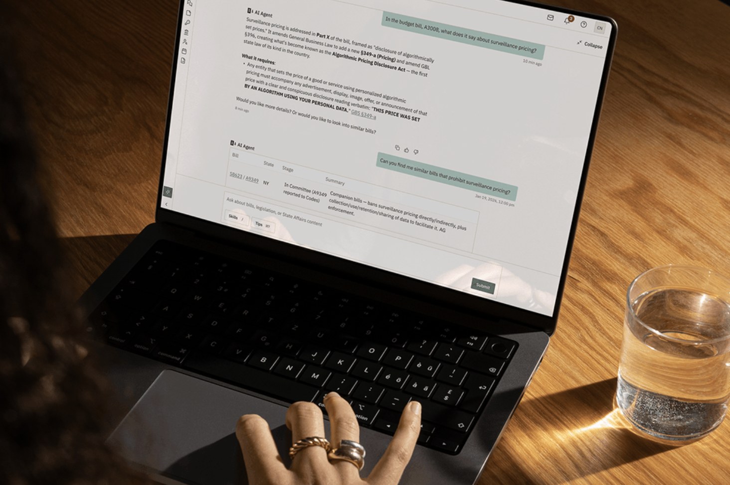 A hand on a MacBook showing the State Affairs AI Agent answering a question about a bill, on a wooden desk